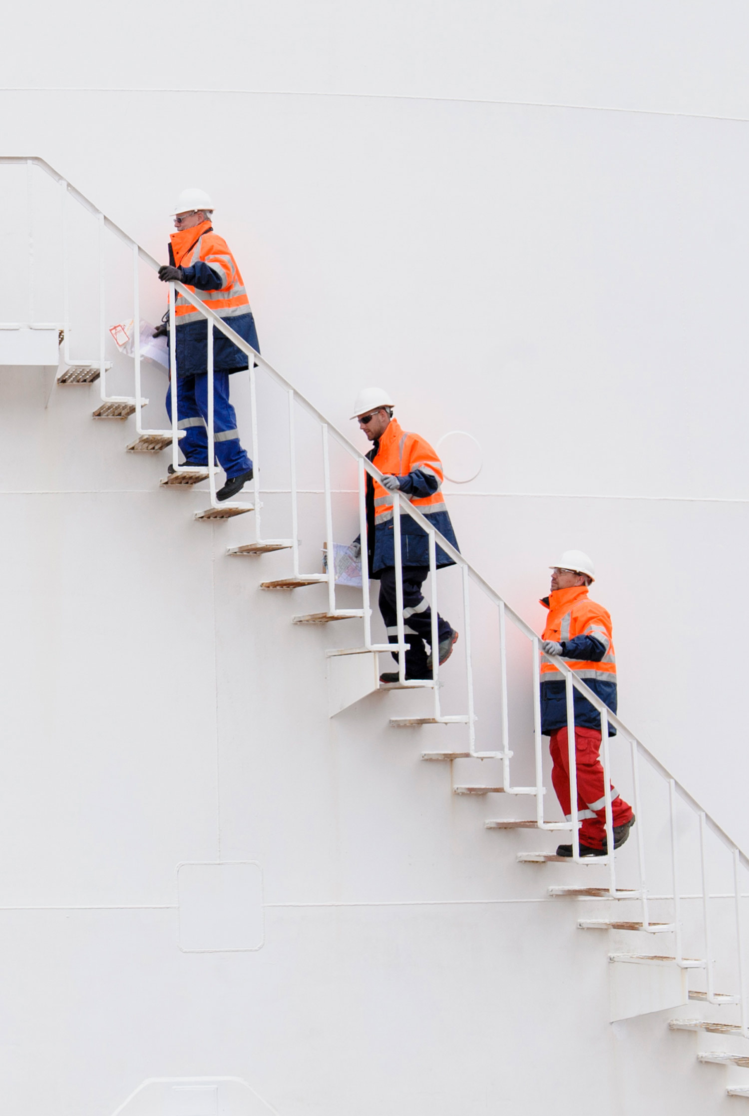 Three Men in hard hats wearing white hardhats and safety coats walking up a white metal staircase